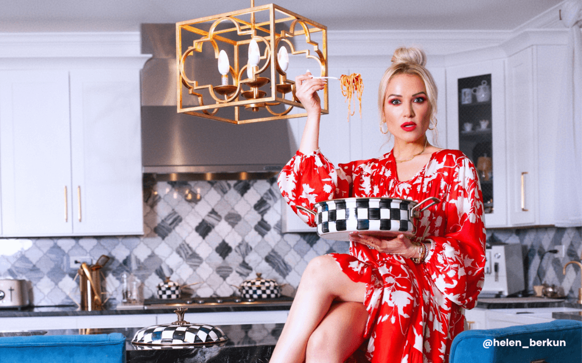 A LTK influencer in red and white printed dress sitting on a kitchen counter eating spaghetti from a black and white checkered bowl.