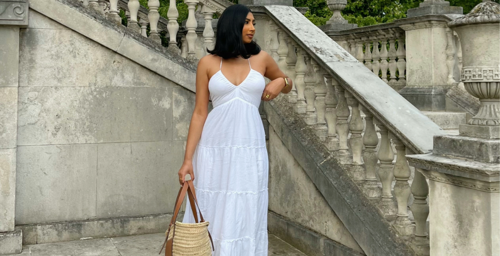 Creator in a white sundress standing by ornate stone stairs, holding a woven tote bag.