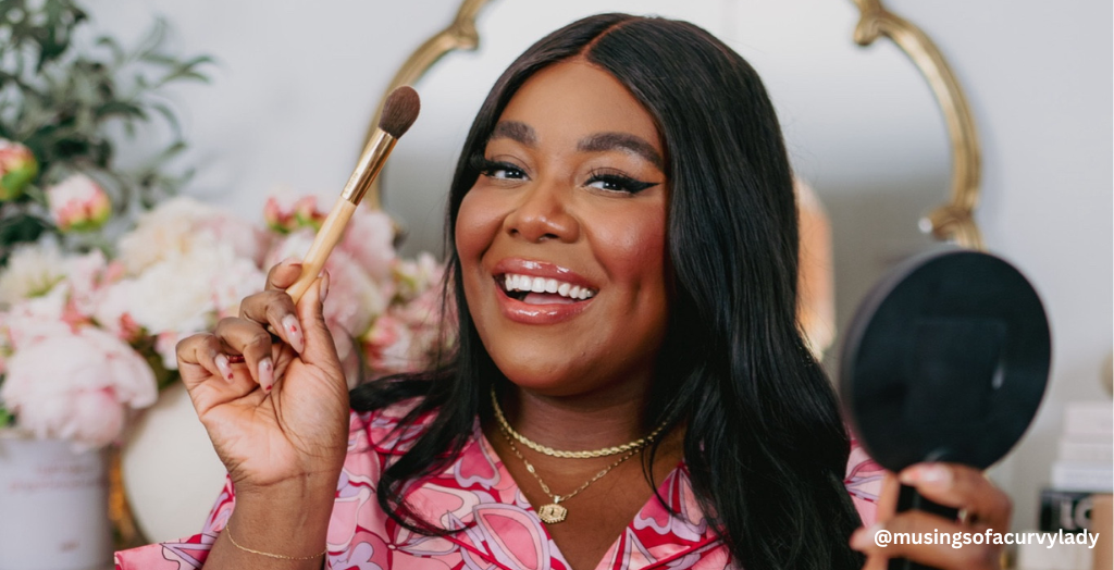 A Creator posing for a photo in front of pink florals while holding a makeup brush and mirror