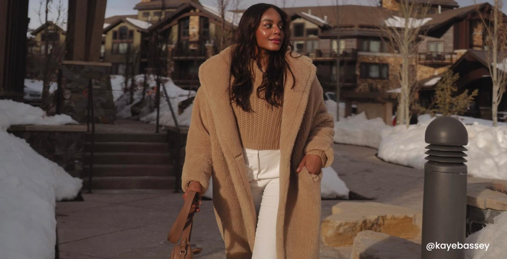 Content Creator @kayebassey in a tan coat and white pants walking through a snowy resort town for a brand collaboration.